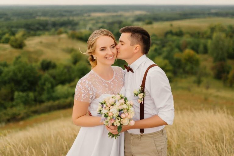 optimize- newlyweds-are-standing-against-beautiful-landscape-with-umbrella-groom-kisses-beautiful-bride-cheek